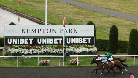 There is a sign saying Kempton Park with Unibet repeated three times underneath it. There are three horses racing on the right side of the image, each with its respective rider. There are are hedges in the background and flowers across the grass.