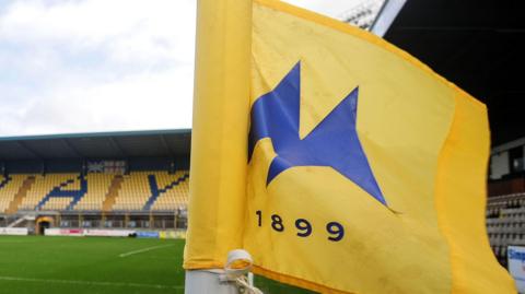 Torquay United's Plainmoor stadium. A yellow and blue flag with "1899" is in the foreground and the pitch and stands in the background.