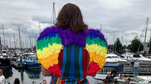A woman stands with her back to the camera, looking across the marina. She is wearing a pair of angel wings on her back. There are lots of boats in the background and a grey, cloudy sky.
