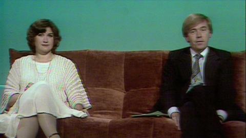 Dame Jenni Murray on the left on a sofa with a white dress on. Bruce Parker on the right in a black suit with a grey tie.