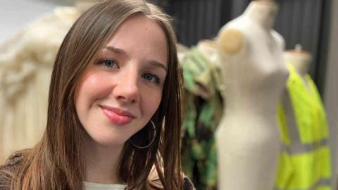 Scarlett Baker has long brown hair and is wearing a textured brown cardigan over a light top. Behind her, there are some mannequins, one features a cream‑coloured dress, another has a green patterned outfit, and a third is dressed in a bright yellow high‑visibility jacket but they are not in focus. 