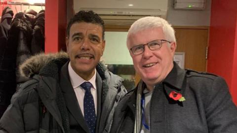 Nick Waites is pictured with former footballer Chris Kamara. Nick has short white hair and wears glasses. He is on the right of the picture. To his left is Chris Kamara. He has short black hair and a thing black moustache. Both are smartly dressed and wearing coats.