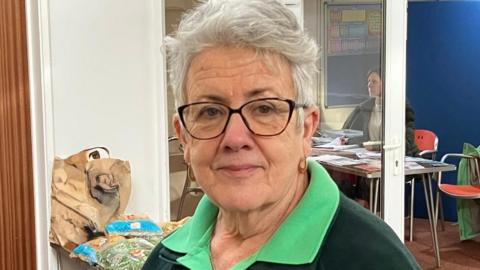 A woman in a green polo top and black jacket with short grey hair and glasses with produce behind her. 