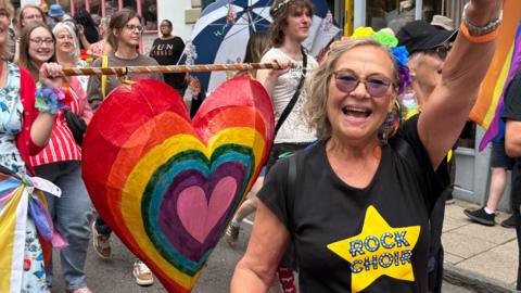 A smiling woman in sunglasses, flower crown and pink tutu waves in front of a rainbow coloured heart