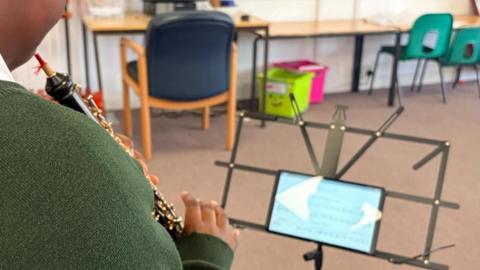 A child wearing glasses playing a clarinet in a classroom, reading sheet music from a stand with computers and chairs visible in the background.
