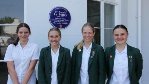 Four girls in year 9 and 10 stand in front of a white external wall with glass windows. There is a blue plaque on the wall behind them reading 'Dr Amelia Le Pelley' in white writing. Three of the girls have a forest green blazer on, all are wearing white shirts and smiling at the camera. 