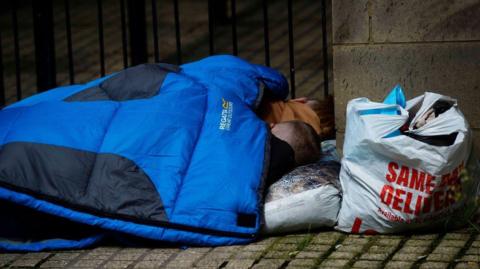 Two people lie on a street corner in a blue sleeping bag. A plastic bag is near to them.