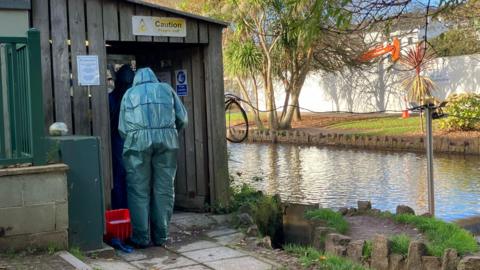 A person is seen in a hazmat suit looking into a wooden storage shed which is situated along a canal.