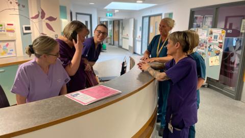 Three women in scrubs behind a nurses station desk and three women in scrubs leaning on the desk on the other side