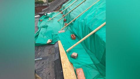 A roof without slates or tiles left with green sheets held down with bricks and planks of wood