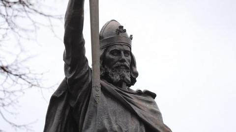 A statue of King Alfred the Great. He is wearing a helmet and holding a sword in the air. 