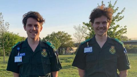Ryan Appleton stands in a grass field alongside his brother. They both wear matching ambulance green uniforms. They both have brown hair and are smiling at the camera.