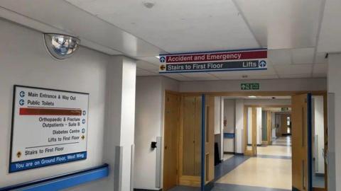 A hospital corridor which has a series of open wooden doors and a sign for Accident and Emergency hanging from the ceiling.