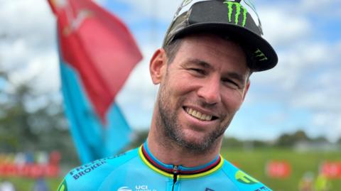 Mark Cavendish, a man with short brown hair and stubble wearing a turquoise cycling top and black cap. There is a Manx flag in the background.