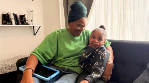 Zaeah and Grandma Joann are both sat on a sofa. Joann is smiling with Zaeah tablet on her lap. Zaeah is smiling with her hands covering her mouth. 