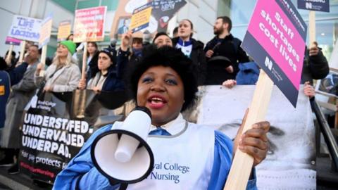A nurse, holding a megaphone in one hand and a Royal College of Nursing placard in the other, stands on a picket line outside UCH hospital in London in 2023. She is standing in front of other pickets who are holding brightly coloured banners. 