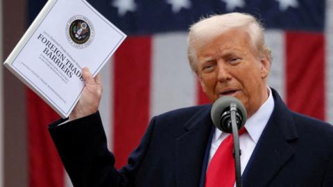 Donald Trump holds a document that says 'Foreign Trade Barriers'. He wears a blue suit, white shirt and red tie. There is a mic in front of him and a US flag behind.