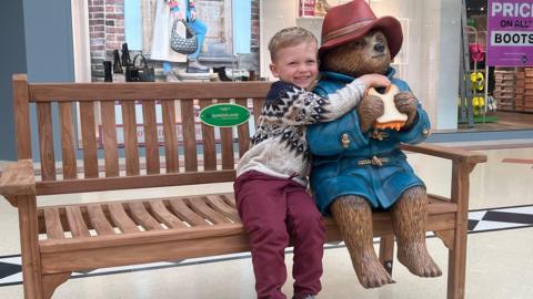 Three-year-old Freddie, sits next to and hugs the newly unveiled statue of Paddington at the Bridges shopping centre in Sunderland. Both Freddie and Paddington are seated on a wooden park bench.