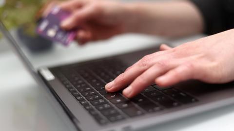 A person types on a grey laptop with a black keyboard while holding a purple debit card.