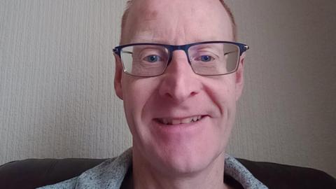 Alan Mcaulay is smiling at the camera. He is wearing glasses that have dark rims at the top. There is white wallpaper behind him