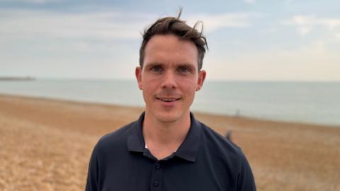 Sam Johnson who has brown short hair and a black polo top. Sam is standing on Brighton beach which has a shingle bank