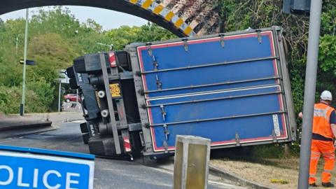 A large blue lorry that has overturned in front of a railway bridge. Surrounding cars are stationary.