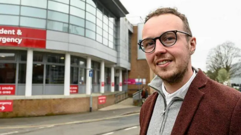 A man in a grey top and a brown coat stands outside a hospital