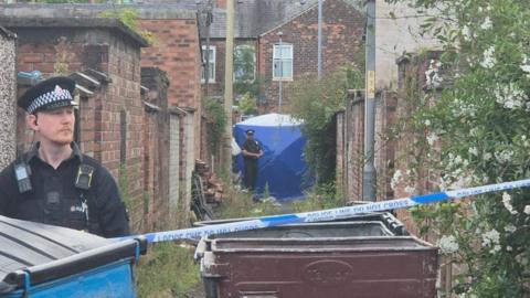 A wide shot of an alleyway where a police cordon has been set up. A blue forensic tent has been erected and is being guarded by two police officers in uniform. 