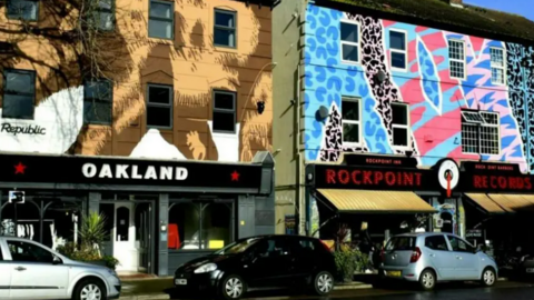 A general view of Rockpoint Records, a bar in New Brighton, Wirral. The walls of the building are painted in vivid colours. An awning hangs down above tables and chairs outside, and cars line the street. 