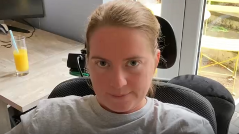A woman with tied back brown hair wearing a grey top sits in a black padded wheelchair.