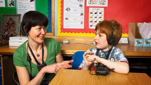 A deaf school boy in a school class room using sign language. An seven year old red head boy fitted with a voice amplifier communicates with his teacher using sign language.