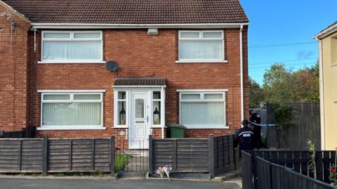 Two floral tributes placed outside the front gate of a red-brick house. Two police officers are at the side of the property. An area is cordoned off with police tape.