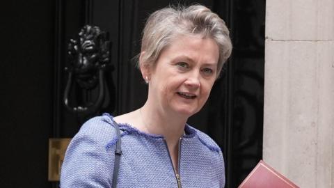 Home Secretary Yvette Cooper looks towards the camera while wearing a blue top with cropped short hair outside Number 10 Downing Street, holding a red file just in view, in London in July.