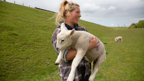 Hollie is wearing a red and black and white checker shirt and holding a lamb 