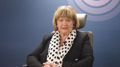 Baroness Heather Hallett sits in the inquiry chair wearing a black suit and spotty scarf, with the UK Covid-19 Inquiry logo on the wall behind her