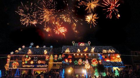 Exterior shot of a house with elaborate Christmas lights and fireworks and a crowd outside 