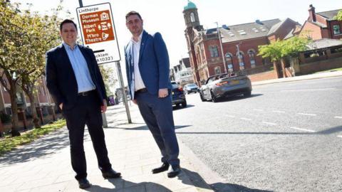 Tees Valley Mayor Ben Houchen and Middlesbrough Mayor Chris Cooke standing on Linthorpe Road where a cycle lane has been removed. There is no longer any trace it ever existed. Houchen wears a black suit with a pale blue shirt and Cooke wears a blue suit with a white shirt. The photo has been taken at a tilted angle.