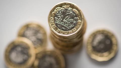 Pound coins are pictured on a white surface. Coins are scattered around and there a few coins stacked up one on top of each other.