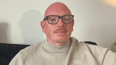 A man with glasses in a beige turtleneck sits on a sofa. 