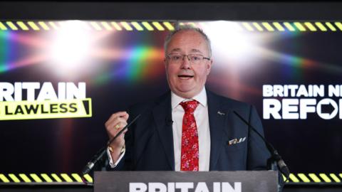 A man in a suit and tie in front a screen saying "Britain is Lawless"