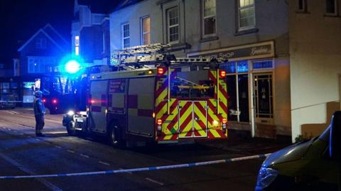 A fire engine outside a burnt barber's shop. It is dark and a blue light flashes. 
