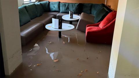 Floodwater covers the floor of a lounge seating area, with a curved dark sofa, a red armchair and two small round tables partially surrounded by standing water and scattered leaves.