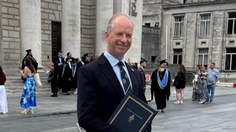 A man is stood in a blue shirt with a black blazer on top. He has a black tie with gold detailing on it. He is holding a certificate. He is smiling looking away from the camera. Behind him is the Guildhall.