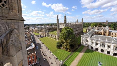 King's College chapel, Cambridge, with a large tree in front. The  chapel has a number of spires along it's length. To the right is a white building known as Senate House. It has 5 arched doorways on the ground floor and the first floor has large windows made up of several smaller panes. The grass in front of both buildings is striped and the sky is blue with a number of white clouds. 