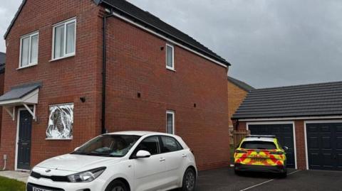Two police vehicles are parked on the drive of a newbuild house. One is a white car without any police markings on. A police officer is sat in the driver's seat. The other car has full police colours. There is a silver foil sheet over the downstairs window of the house.