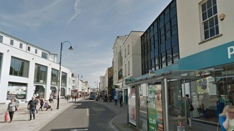 Street view of High St in Cheltenham. The edge of Poundland is visible to the right. A bus has pulled into a stop in the distance, and the street shows lots of people walking up and down.