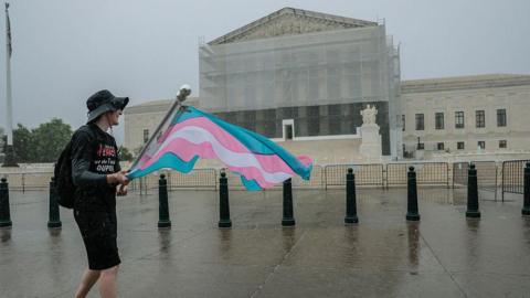 Man in black T-shirt, black shorts and black hat carries flag with blue, pink and white stripes on rainy day with Supreme Court building, wrapped in light scaffolding in the background