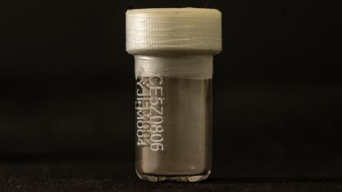 A small clear viall with a white plastic top and tape around the lid. It is lit against a black background. On the front is a code. Inside it looks great and at the bottom there is a coating of dust.