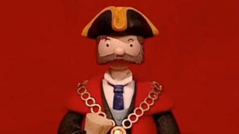 The Mayor from the stop motion children's show, Trumpton. He has a large brown beard and is wearing a black and yellow tricorn hat, a red and black robe and a gold chain. He is standing against a red background