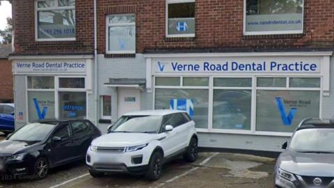 The corner plot of a row of two-storey, red-brick terraced houses. The dentist takes up the bottom floor. The shop front is painted grey with signs that read: Verne Road Dental Practice. There are cars parked in front.